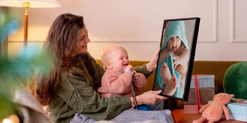 Vrouw met een baby op schoot in een woonkamer, die een gepersonaliseerde wanddecoratie van de baby omhooghoudt terwijl de baby ernaar kijkt en giechelt, ter illustratie van albelli’s ideeën voor fotocadeaus voor de eerste kerst van een baby.