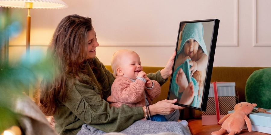 Vrouw met een baby op schoot in een woonkamer, die een gepersonaliseerde wanddecoratie van de baby omhooghoudt terwijl de baby ernaar kijkt en giechelt, ter illustratie van albelli’s ideeën voor fotocadeaus voor de eerste kerst van een baby.