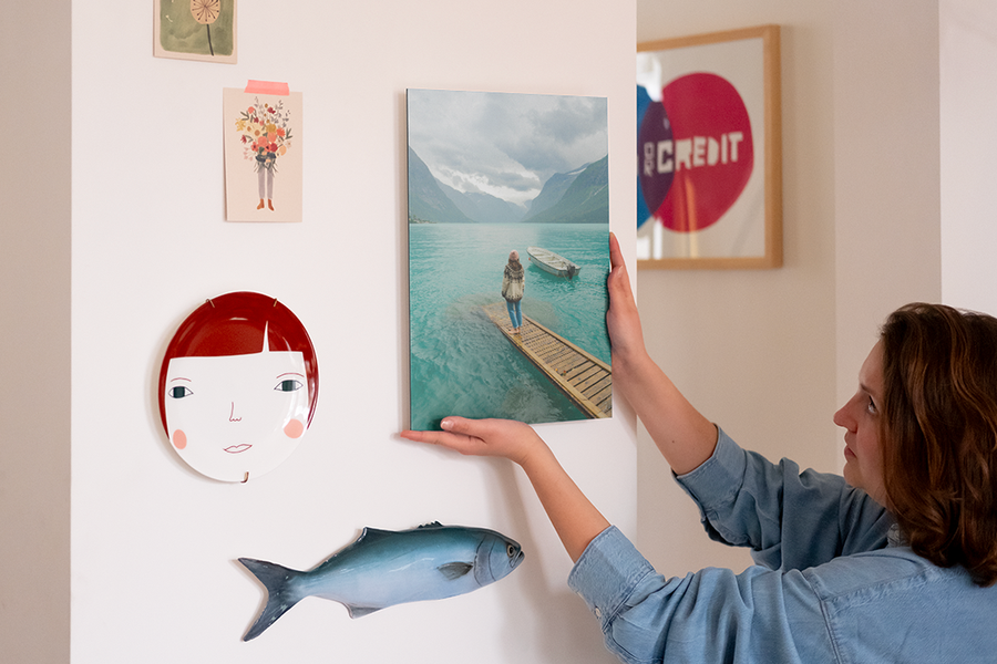 Een vrouw hangt gepersonaliseerde wanddecoratie aan haar muur, naast een decoratief bord en enkele fotoafdrukken, ter illustratie van albelli's laatste stap om je wanddecoratie op te hangen en de look te voltooien.