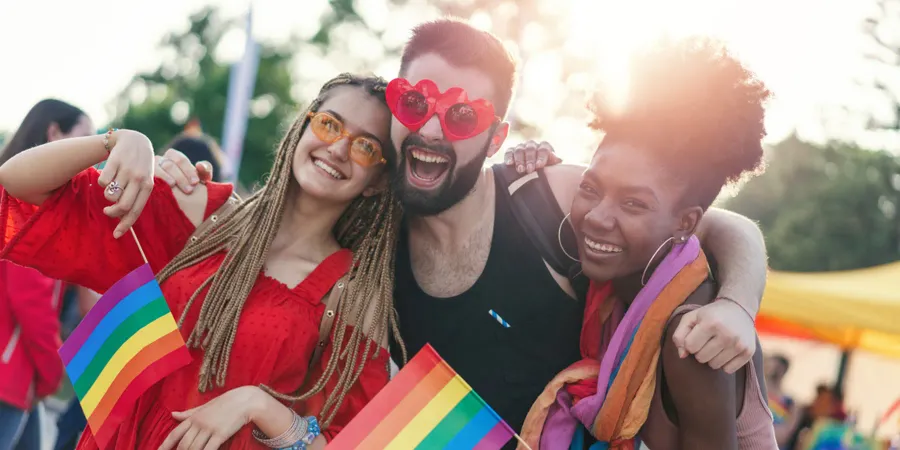 Drie vrienden glimlachen op een Pride-evenement, met regenboogvlaggen in de hand en een zonnebril op, als viering van de vreugde en verbondenheid van LGBTQI+ Pride.