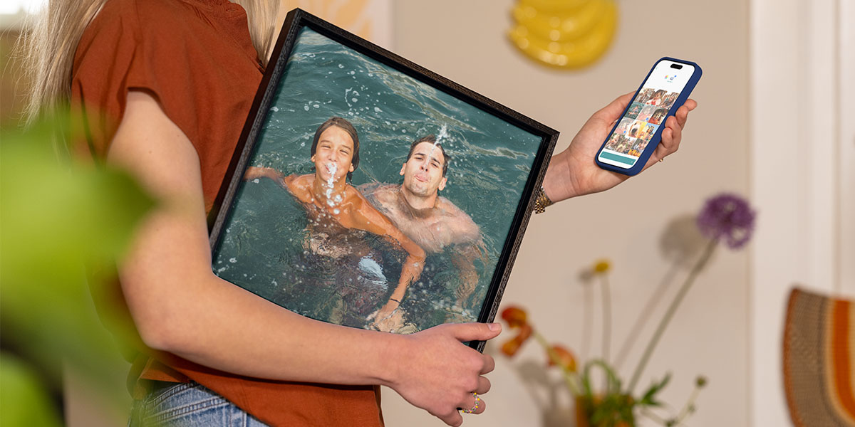 Een vrouw houdt een ingelijste vakantiefoto van een man en een jonge jongen die in het water spelen onder haar arm, terwijl ze op haar telefoon foto's selecteert, ter illustratie van albelli's gids om vakantiefoto's om te zetten in wanddecoratie.