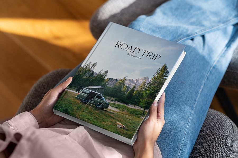 Een albelli koffietafelboek met de titel Road Trip ligt op iemands schoot, met een boslandschap en camper op de omslag, als inspiratie voor een reiskoffietafelboek.