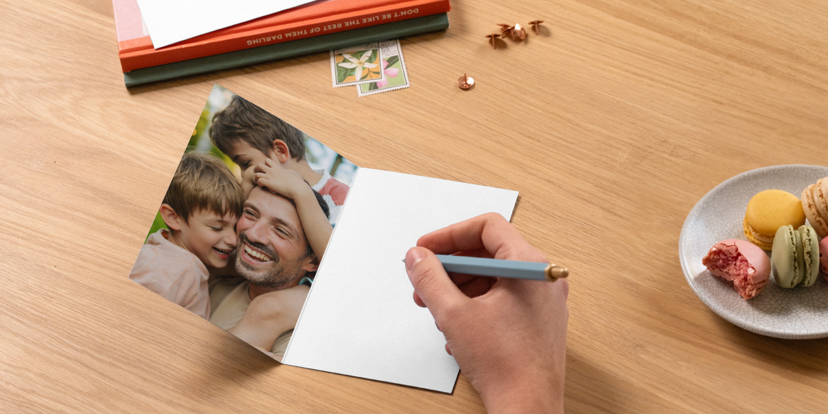 Een open gepersonaliseerde albelli vaderdagkaart toont een vader met zijn twee zoons, met een pen klaar om een boodschap te schrijven naast boeken en een bord macarons, als inspiratie voor wat je op een vaderdagkaart schrijft.