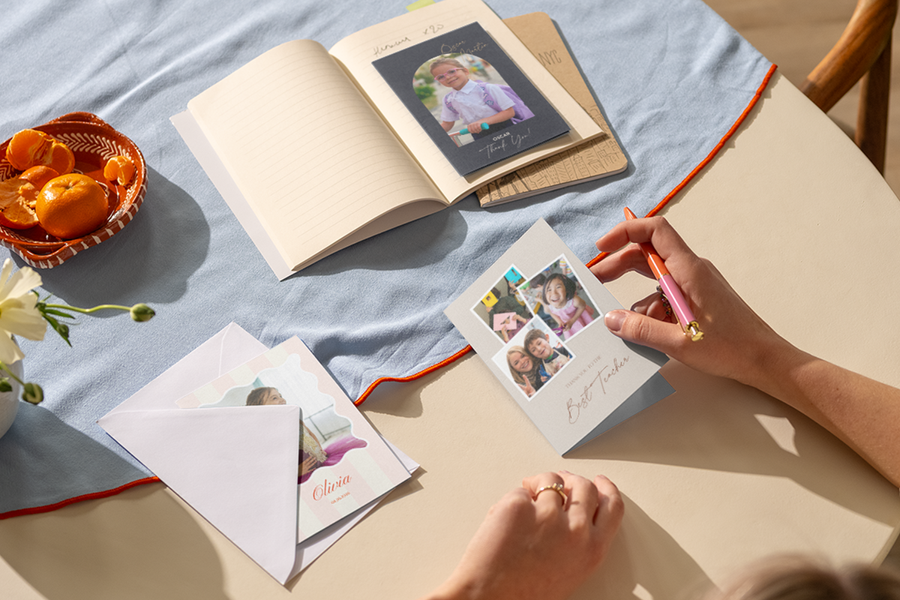 De hand van een vrouw rust op een tafel en staat klaar om te schrijven in een gepersonaliseerd albelli bedankkaartje voor een leraar, met daarop verschillende foto’s van blije kinderen.