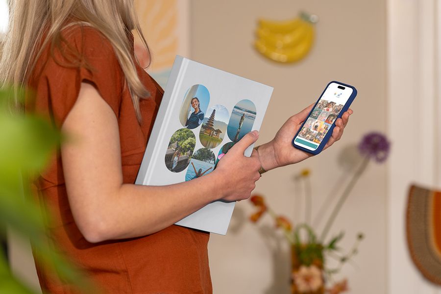 Een vrouw houdt een gesloten albelli vakantie fotoboek vast met familiereisfoto’s op de cover terwijl ze op haar telefoon naar reisfoto’s kijkt, als illustratie van hoe je de juiste foto’s kiest voor een gepersonaliseerd reisfotoboek.