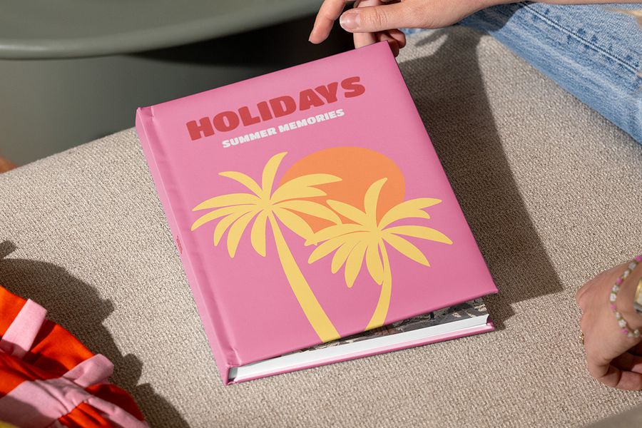 Een gesloten albelli reisfotoboek met de titel “Holidays, summer memories” en geïllustreerde palmbomen op een roze cover wordt vastgehouden door handen die klaar zijn om het open te slaan, als inspiratie voor hoe je een vakantie fotoboek structureert en je reisverhaal vertelt.