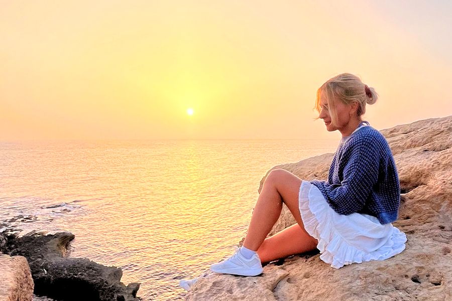 Een vrouw zit op rotsen en kijkt uit over de zee en de zonsondergang, passend bij albelli's tips voor een avontuurlijke vakantie thuis.