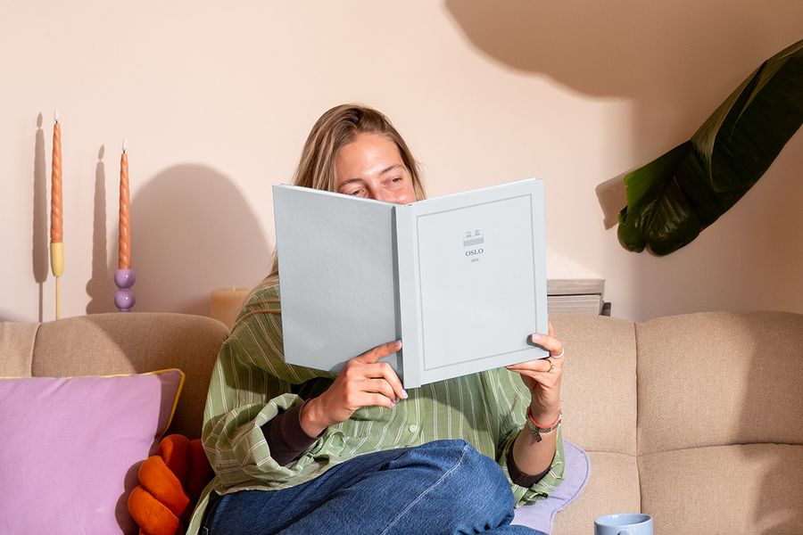 Een vrouw zit op de bank naast een kleurrijk kussen en bekijkt een gepersonaliseerd reisfotoboek met de titel 'Oslo', ter illustratie van albelli's nieuwe Summer Travel Series ontwerpen.