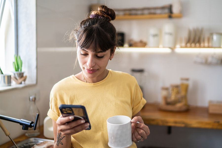 Een vrouw in haar keuken houdt een mok vast terwijl ze op haar telefoon kijkt, ter illustratie van albelli's tips om fotogewoontes op te bouwen die je ook echt volhoudt.