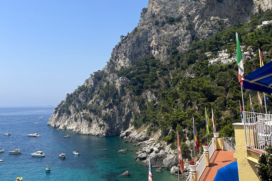 Een mediterrane kustlijn met heuvels bij een blauwe zee bezaaid met bootjes, gezien vanaf een balkon op de voorgrond, als beeld van de charme van een septembervakantie in Europa.