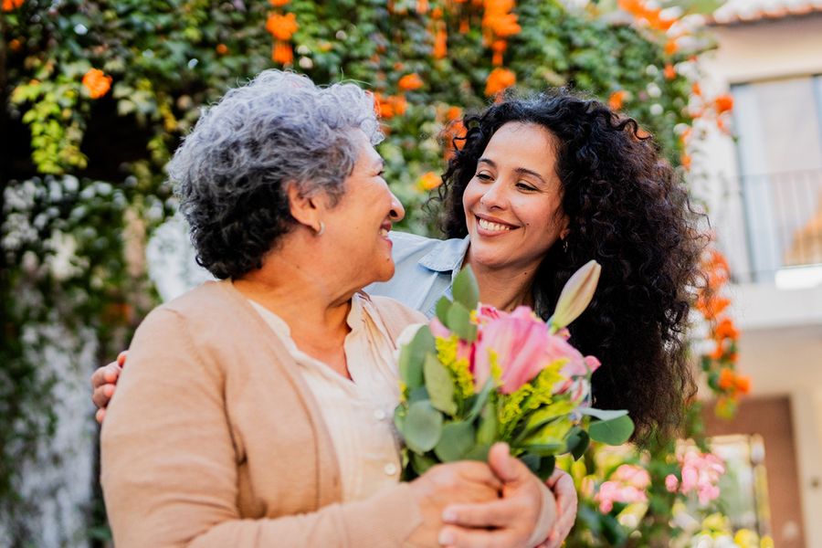 Een jonge vrouw glimlacht naar haar moeder, die bloemen in haar handen houdt, ter illustratie van albelli's ideeën voor wat je kunt schrijven in een huwelijksjubileumkaart voor je ouders.