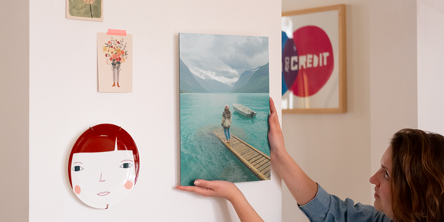 Een vrouw hangt gepersonaliseerde wanddecoratie aan haar muur, naast een decoratief bord en enkele fotoafdrukken, ter introductie van albelli's stap-voor-stap gids om wanddecoratie op te hangen met hangkits.