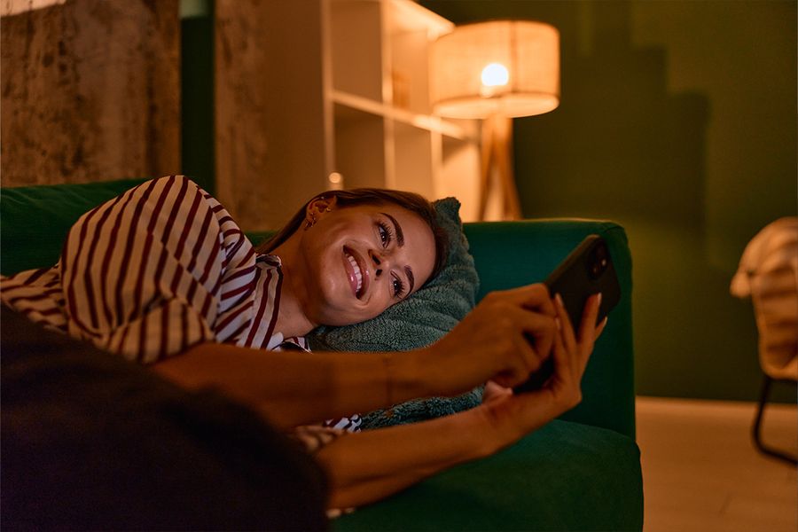 Een vrouw ligt op een donkergroene bank in de woonkamer en kijkt op haar telefoon, verlicht door een lamp voor gezellig licht, ter illustratie van albelli's tips voor hygge-verlichting in huis.