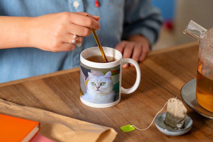 Iemand roert met een lepel in een gepersonaliseerde fotomok met een foto van een witte kat terwijl hij een kopje thee zet, ter illustratie van albelli's tip om een hygge-leeshoek af te maken met een gepersonaliseerde mok.