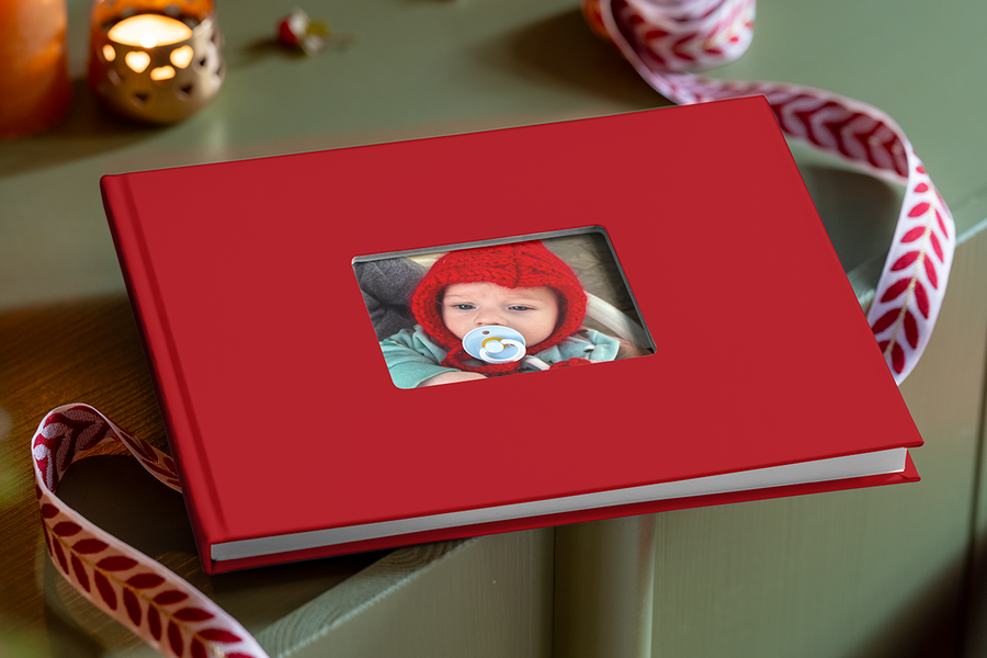 Rood fotoboek met een uitgesneden venster in de cover, waardoor een pasgeboren baby met een rode gebreide muts en een fopspeen te zien is, ter illustratie van albelli’s tips om een fotoboek te maken met de eerste herinneringen van een baby.