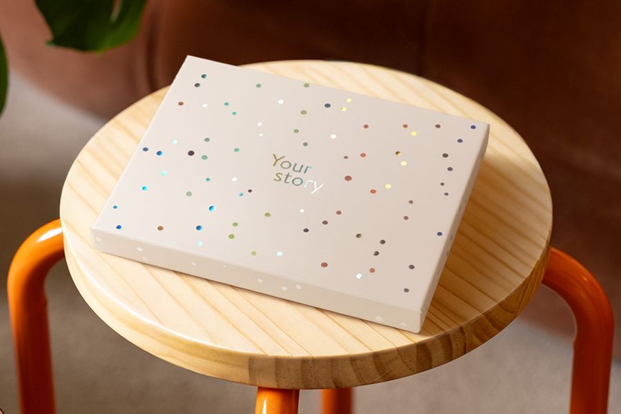Een albelli presentatiedoos staat op een houten kruk, versierd met zilveren holografische cirkels en de tekst 'Your Story', ter illustratie van het matte ivoorkleurige ontwerp met een vleugje holografische glans.