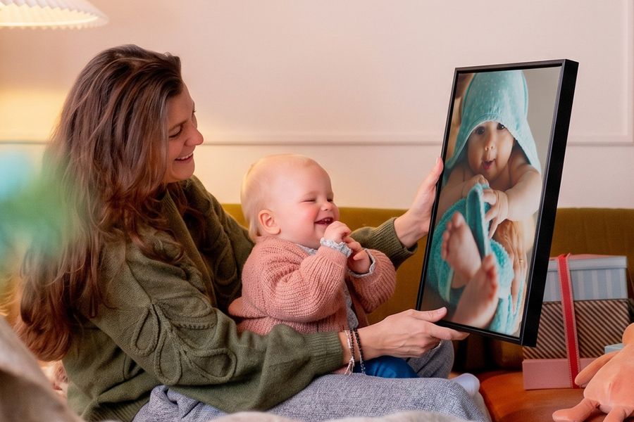 Vrouw met een baby op schoot in een woonkamer, die een gepersonaliseerde wanddecoratie van de baby omhooghoudt terwijl de baby ernaar kijkt en giechelt, ter illustratie van albelli’s ideeën om van babyfoto’s unieke wanddecoratie te maken voor de eerste kerst van een baby.