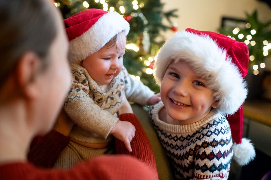 Vrouw met een baby in haar armen naast een lachend jongetje bij een kerstboom, waarbij de baby en het jongetje allebei een kerstmuts en een gebreide wintertrui met patroon dragen, ter illustratie van albelli’s tips om onvergetelijke kerstfoto’s van het gezin en de baby te maken.