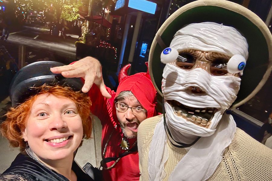 Drie volwassenen in Halloween-verkleedkleding op straat, één met een skeletmasker met uitpuilende ogen, één verkleed als duivel en een vrouw met een zwarte hoed, ter illustratie van albelli's Halloween-teksten voor kostuums.