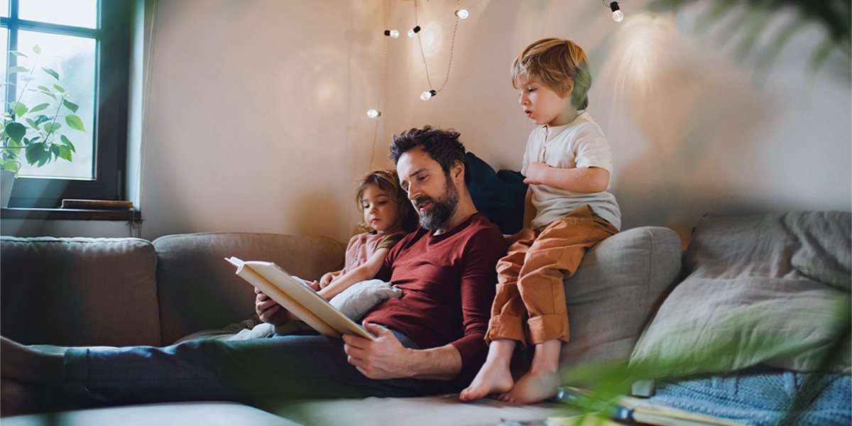 Een vader leest een boek voor aan een jongetje en een meisje op de bank, met warme lichtslingers voor hen, ter introductie van albelli's tips voor een gezellige hygge-herfstdecoratie.