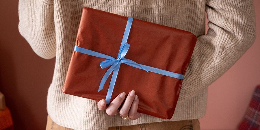 Vrouw die een cadeau verpakt in bruin papier met een blauw lint achter haar rug houdt, ter illustratie van albelli’s checklist om een kerstcadeau te kiezen met een gepersonaliseerd fotocadeau.