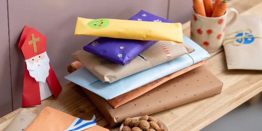 Opgestapelde cadeaus in geel, paars, kraftpapier en papier met stippen, naast een papieren Sinterklaasfiguurtje en een schaaltje pepernoten, ter illustratie van albelli’s gids met de 5 beste gepersonaliseerde Sinterklaascadeaus 2026.