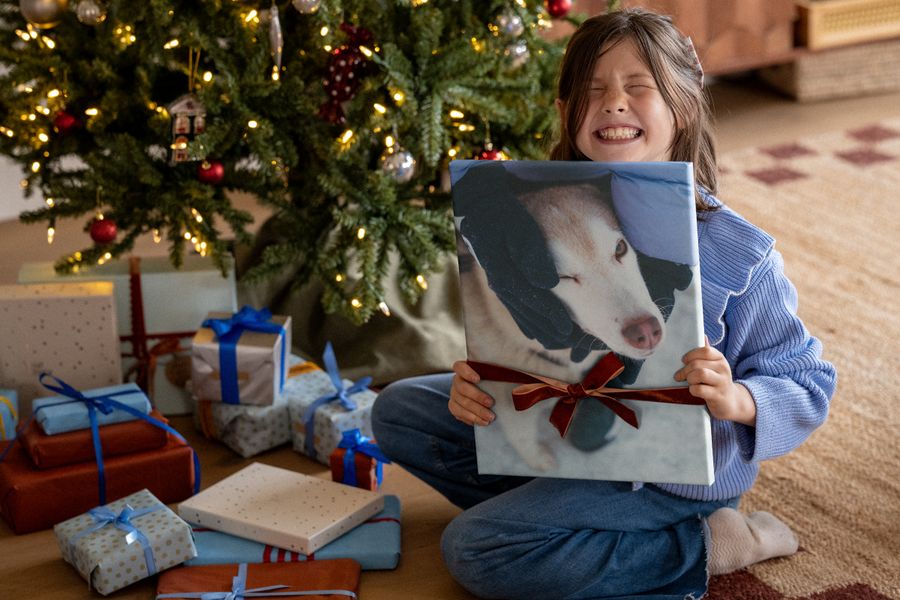 Een lachend meisje in een blauwe trui houdt een canvasprint van een hond met een rood lint omhoog, voor een kerstboom, ter illustratie van albelli’s wanddecoratie als Black Friday-kerstcadeau.