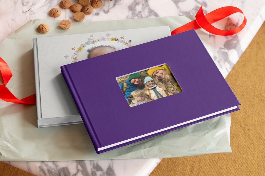 Een paars hardcover fotoboek met een uitsnede in de kaft waarin een gezin van vier met gebreide mutsen buiten te zien is, op een zilvergrijs fotoboek met een krans van geperste bloemen, naast pepernoten en een rood lint, ter illustratie van albelli’s themafotoboeken voor het hele gezin.