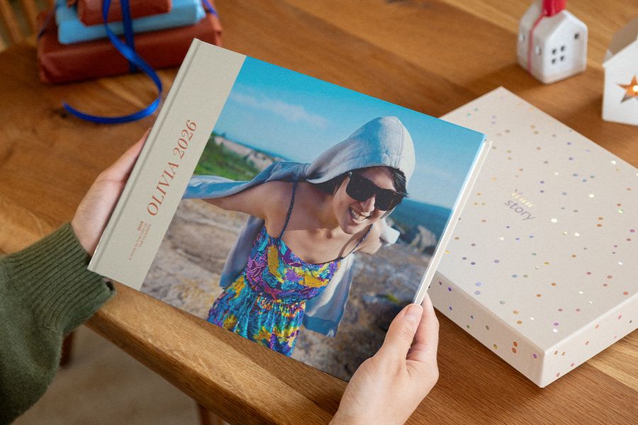 Twee handen houden een fotoboek vast met 2026 op de omslag, waarop een lachende vrouw met zonnebril op een rotsachtige kust staat, naast een presentatiedoos met confetti-stippen, ter illustratie van albelli’s gepersonaliseerde fotoboeken.