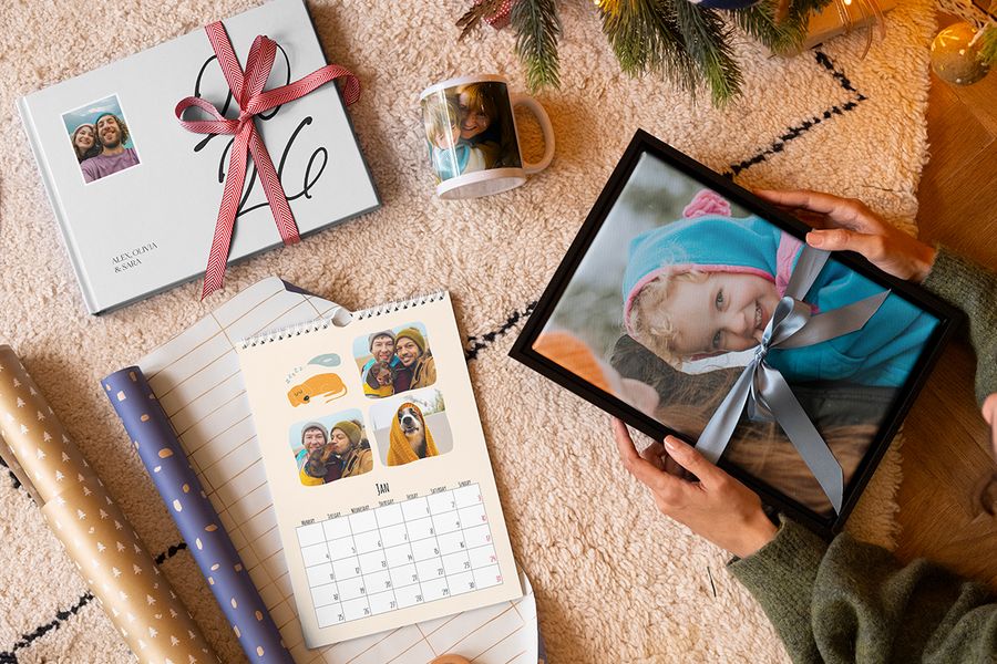 Selectie gepersonaliseerde albelli-cadeaus, waaronder wanddecoratie van een jong meisje met een blauw lint eromheen, een mok met een moeder en kind, een gepersonaliseerde fotokalender en een fotoboek 2026 met een rood lint eromheen, naast inpakpapier en met kerstboomtakken op de achtergrond, ter illustratie van albelli’s tips om de juiste herinneringen vast te leggen in een gepersonaliseerd kerstcadeau.