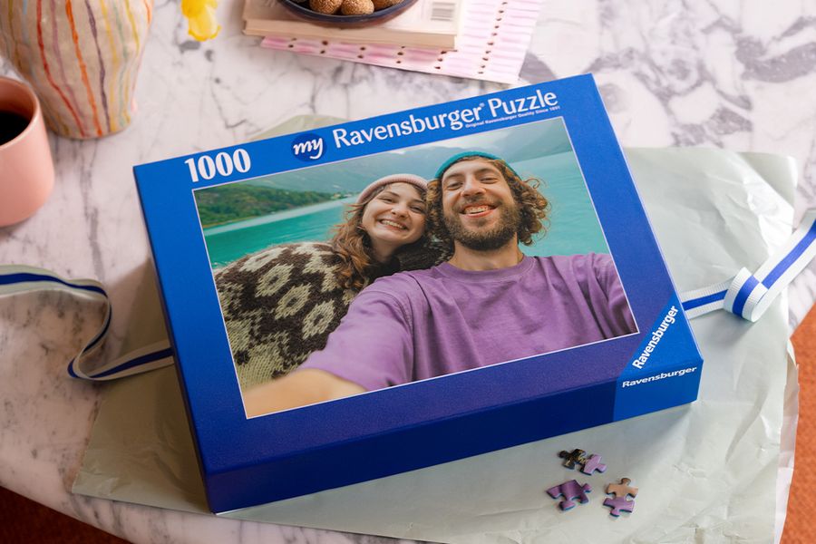 Een blauwe doos van ‘my Ravensburger Puzzle’ met ‘1000’ erop, met een foto van een lachend stel met gebreide mutsen bij een turquoise meer en losse puzzelstukjes ernaast, ter illustratie van albelli’s fotopuzzel als Sinterklaascadeau voor het hele gezin.