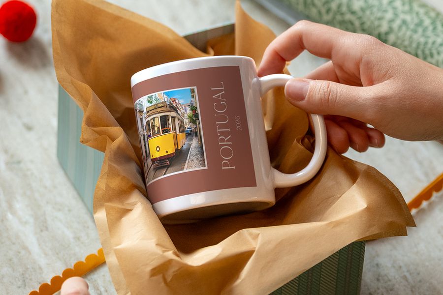 Een hand tilt een mok in kraftpapier op, bedrukt met een gele tram en de tekst ‘Portugal 2026’, ter illustratie van albelli’s gepersonaliseerde mokken als aanwijzing bij een ervaringscadeau.