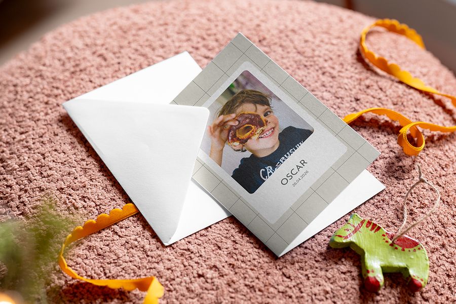 Een vierkante grijsbeige wenskaart met een rasterrand en een foto van een lachend kind dat een donut voor één oog houdt, met de naam ‘OSCAR’ en de datum ‘26.04.2026’ eronder, naast een witte envelop en een oranje lint, ter illustratie van albelli’s tip om het Sinterklaasgedicht te combineren met een persoonlijke kaart.