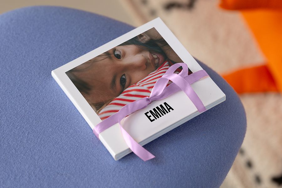 Een klein vierkant fotoboek met een baby in een rood-wit gestreept shirtje op de kaft, met een lila satijnen lint eromheen en de naam ‘EMMA’ in vette hoofdletters, op een blauwe kruk, ter illustratie van albelli’s advies over kleine fotoboeken voor baby’s en peuters.