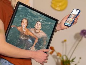 Een vrouw houdt haar telefoon vast met de albelli editor in beeld, terwijl ze onder haar arm vakantiewanddecoratie draagt met daarop twee mannen die plezier hebben in een zwembad, als inspiratie om vakantiefoto’s om te zetten in prachtige wanddecoratie.